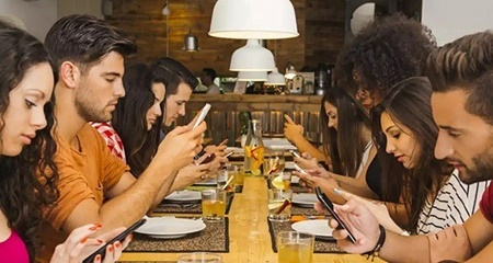 Grupo de jóvenes en una mesa de restaurante, todos mirando sus teléfonos móviles en lugar de conversar entre sí — influencia de las redes sociales y la tecnología en las relaciones